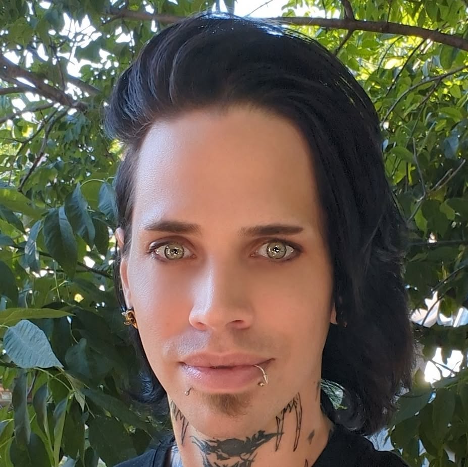 Person with black hair, piercing, and tattoos, standing outdoors with green foliage in background.
