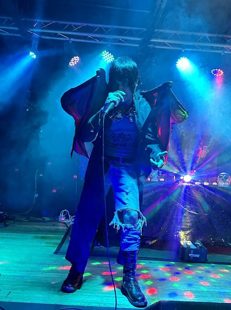 Person with long hair wearing a dark coat and ripped jeans performing on a stage with colorful lights.