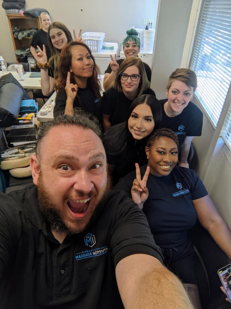 Group of nine people in an office, smiling and making peace signs, with a window and shelves in the background.