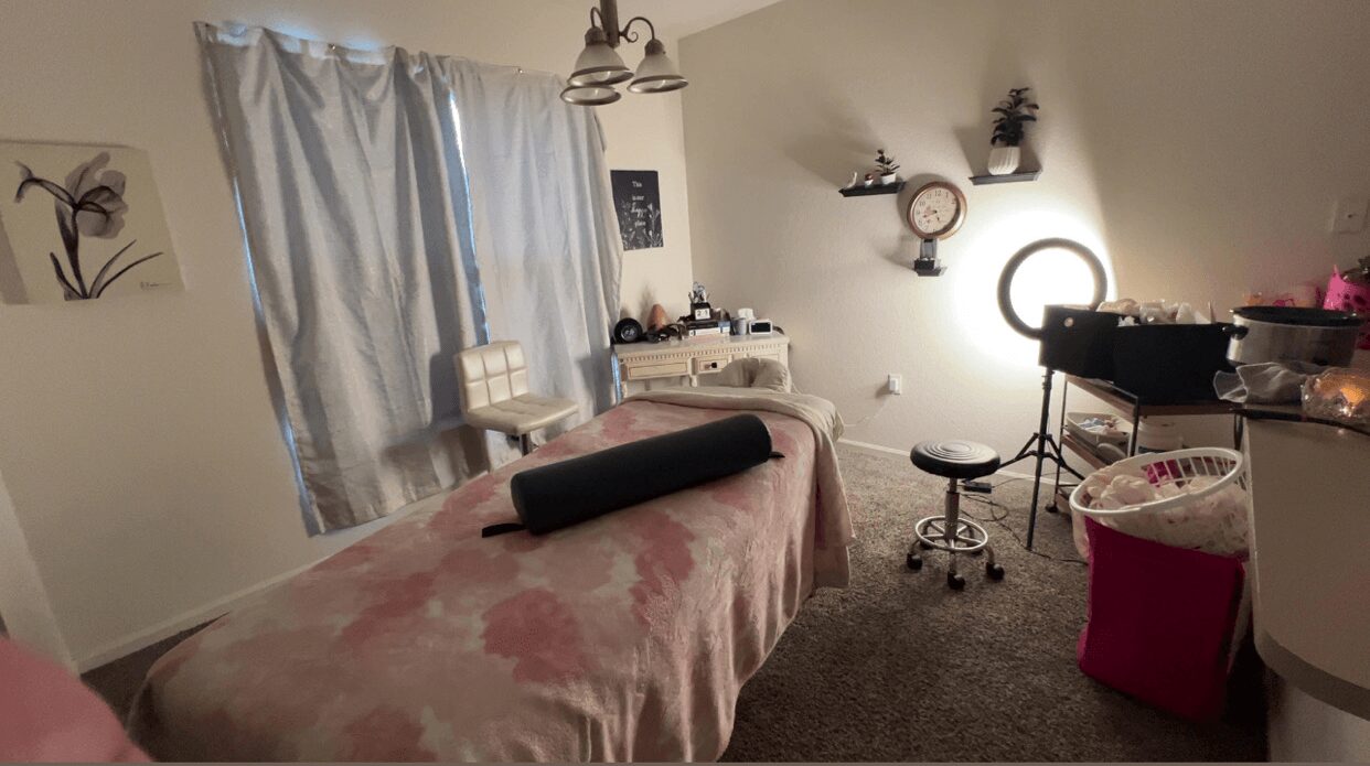 Room with a massage table, curtains, wall decorations, and a small stool, with a pink laundry basket nearby.