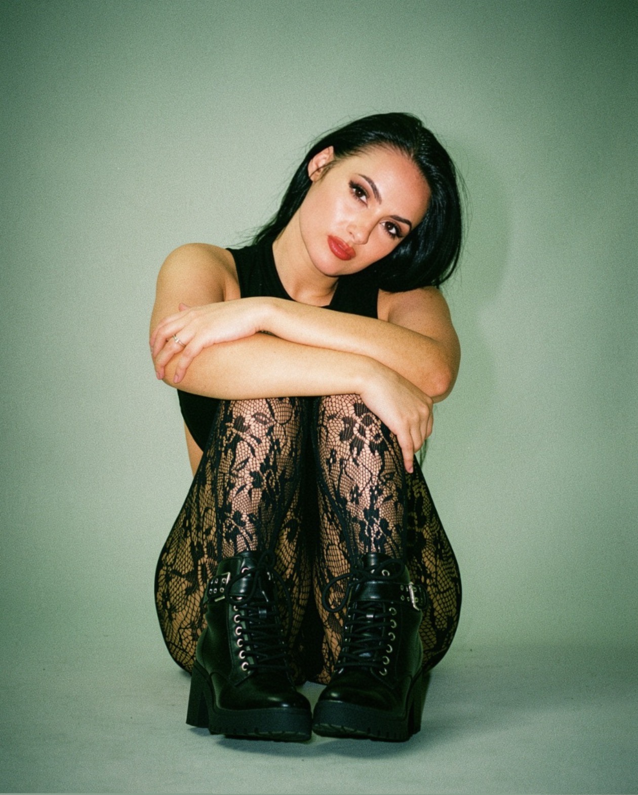 Woman with long dark hair sitting with arms around knees, wearing lace tights and black boots, against a green background.