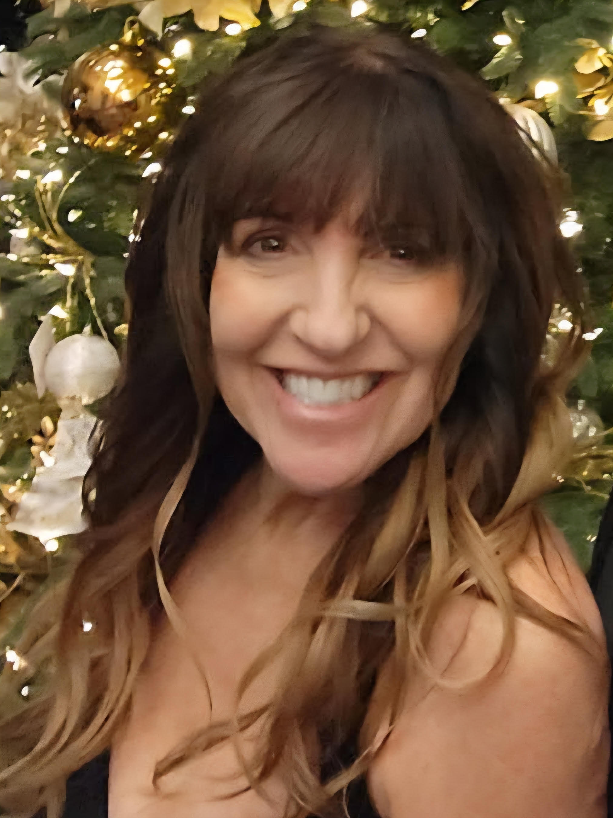 Smiling woman with long wavy hair in front of decorated Christmas tree with gold and white ornaments.
