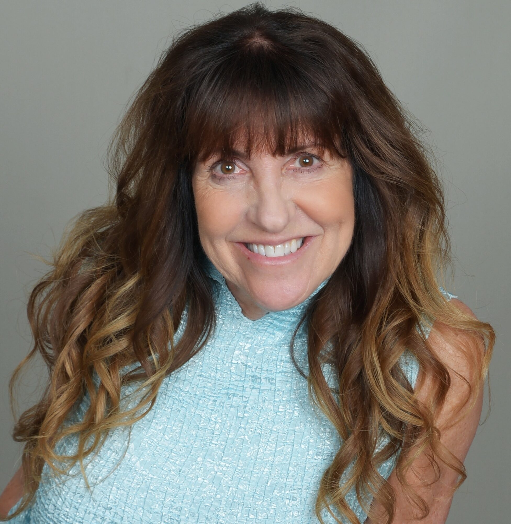 Woman with long wavy brown hair smiling, wearing a light blue top, against a plain background.