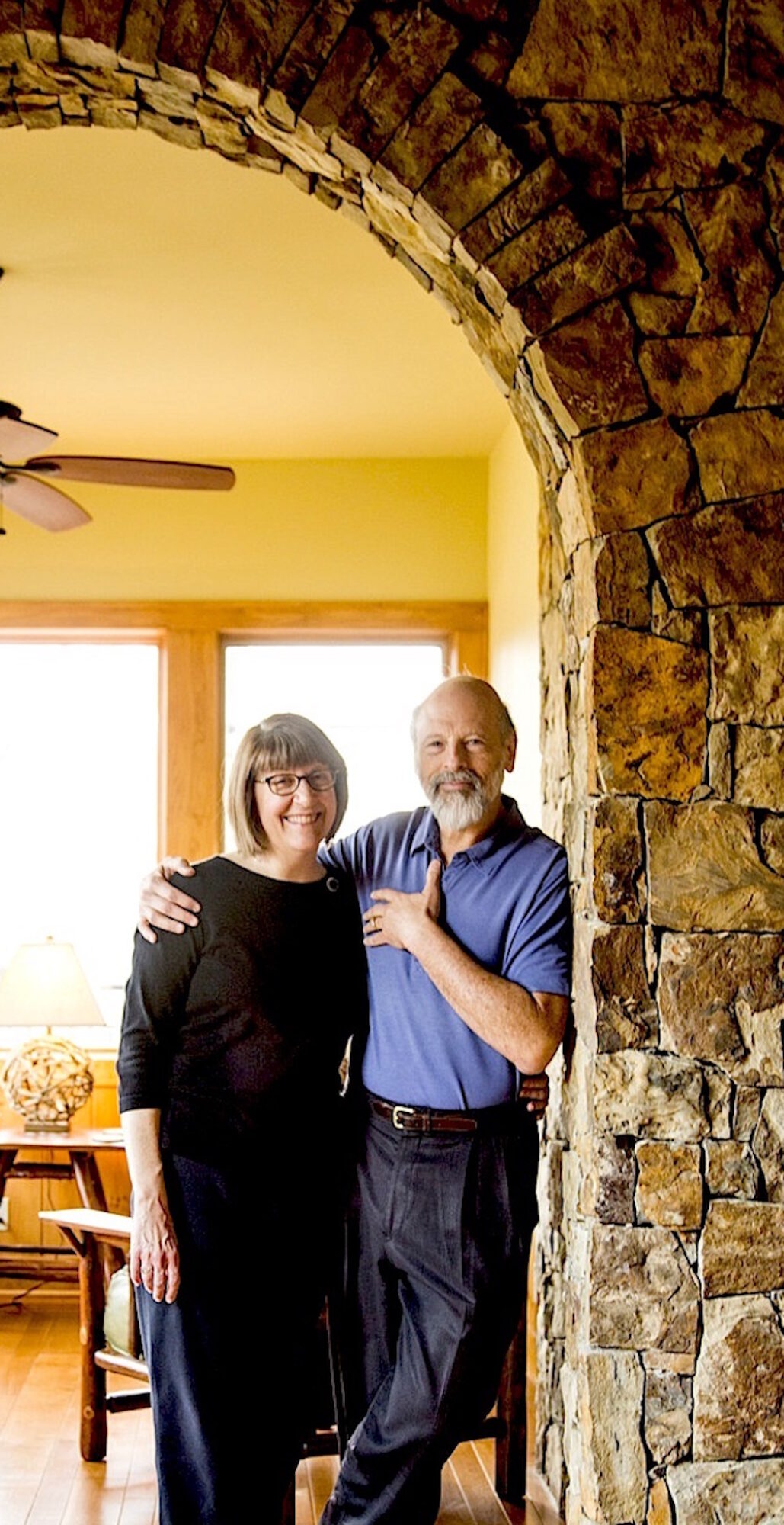 Two people standing together in a room with a stone archway, smiling, with a window and ceiling fan in the background.