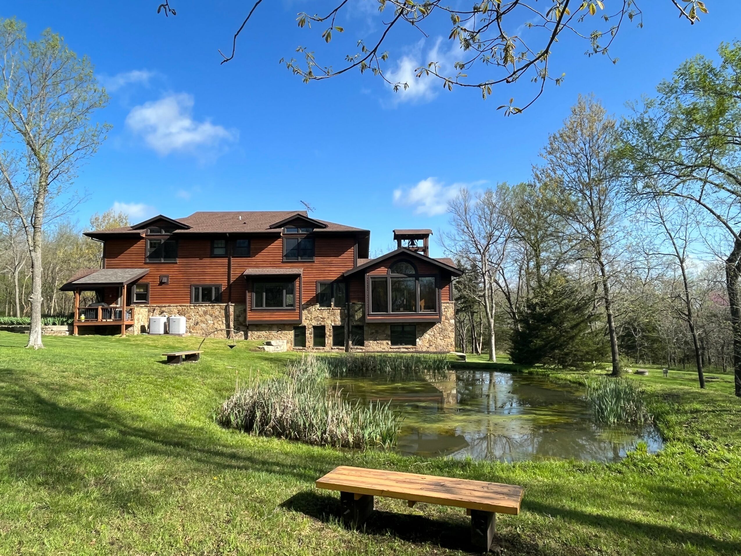 Large wooden house with multiple levels and decks, surrounded by trees, near a pond with a wooden bench nearby.