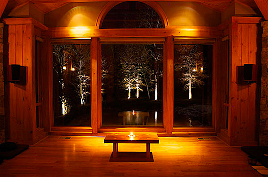 View through large window showing illuminated trees outside at night, warm interior with wooden walls and floor, small table in front.