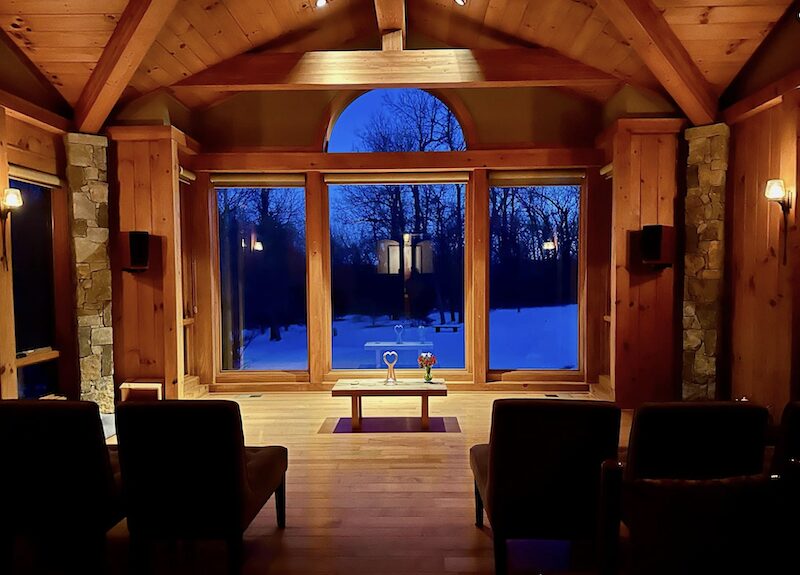 Interior view of a wooden room with large window showing snowy landscape and trees outside, two chairs facing the window, and a small table.
