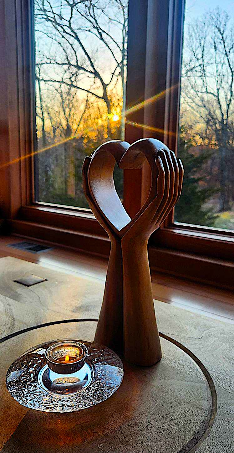Wooden hand sculpture forming a heart shape on a windowsill with sunlight shining through trees outside, candle on table.