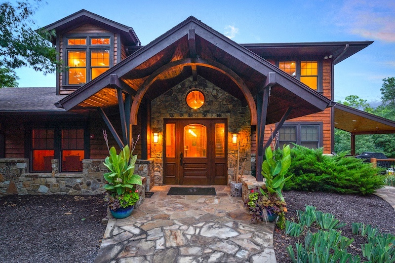 Front view of a house with a stone pathway, wooden exterior, and illuminated windows, surrounded by greenery.