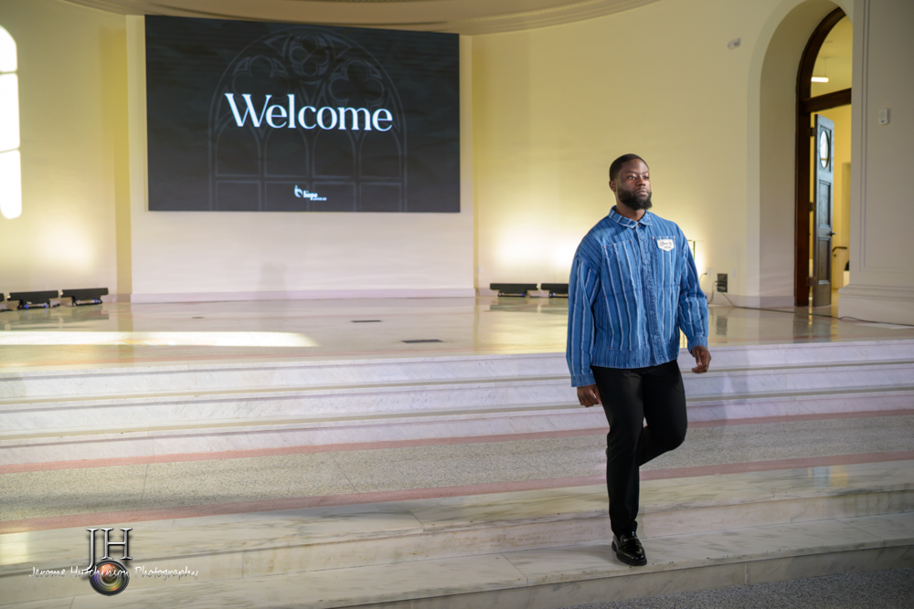 Man walking in front of a large screen displaying 'Welcome' in a spacious room with stairs and arched doorway
