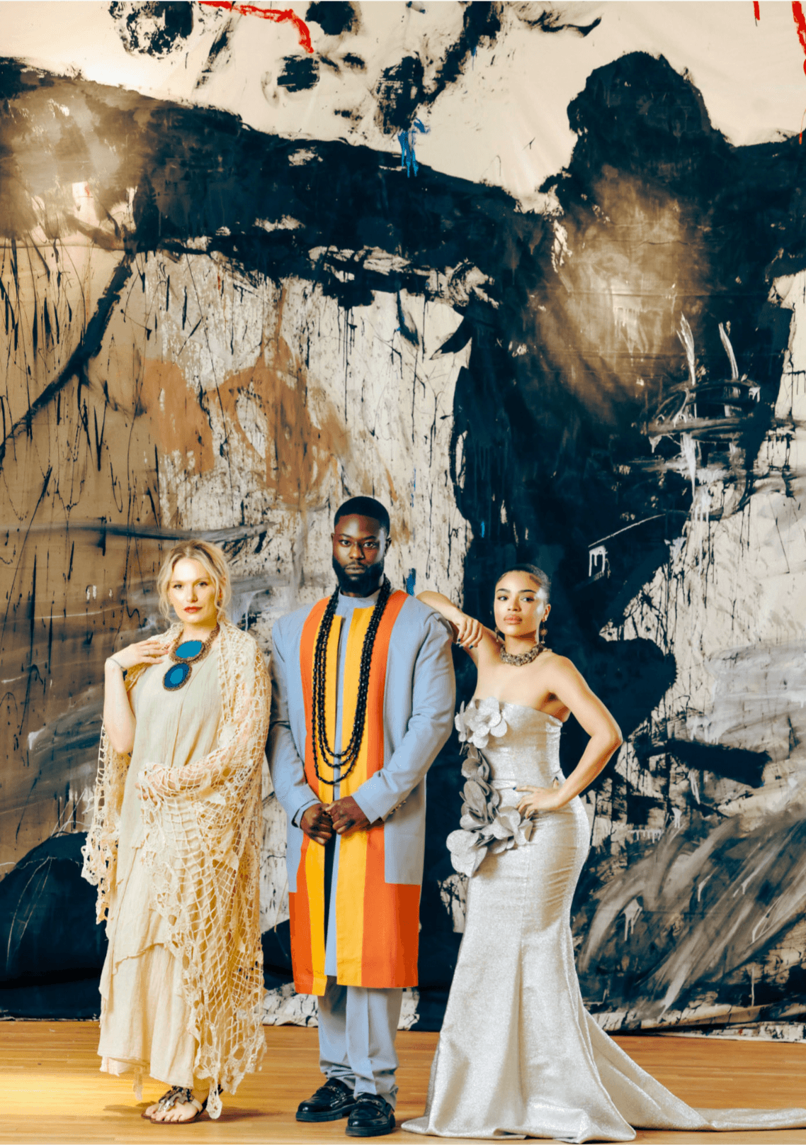 Three people stand in front of an abstract black and white mural, dressed in elegant clothing, with a wooden floor beneath them.