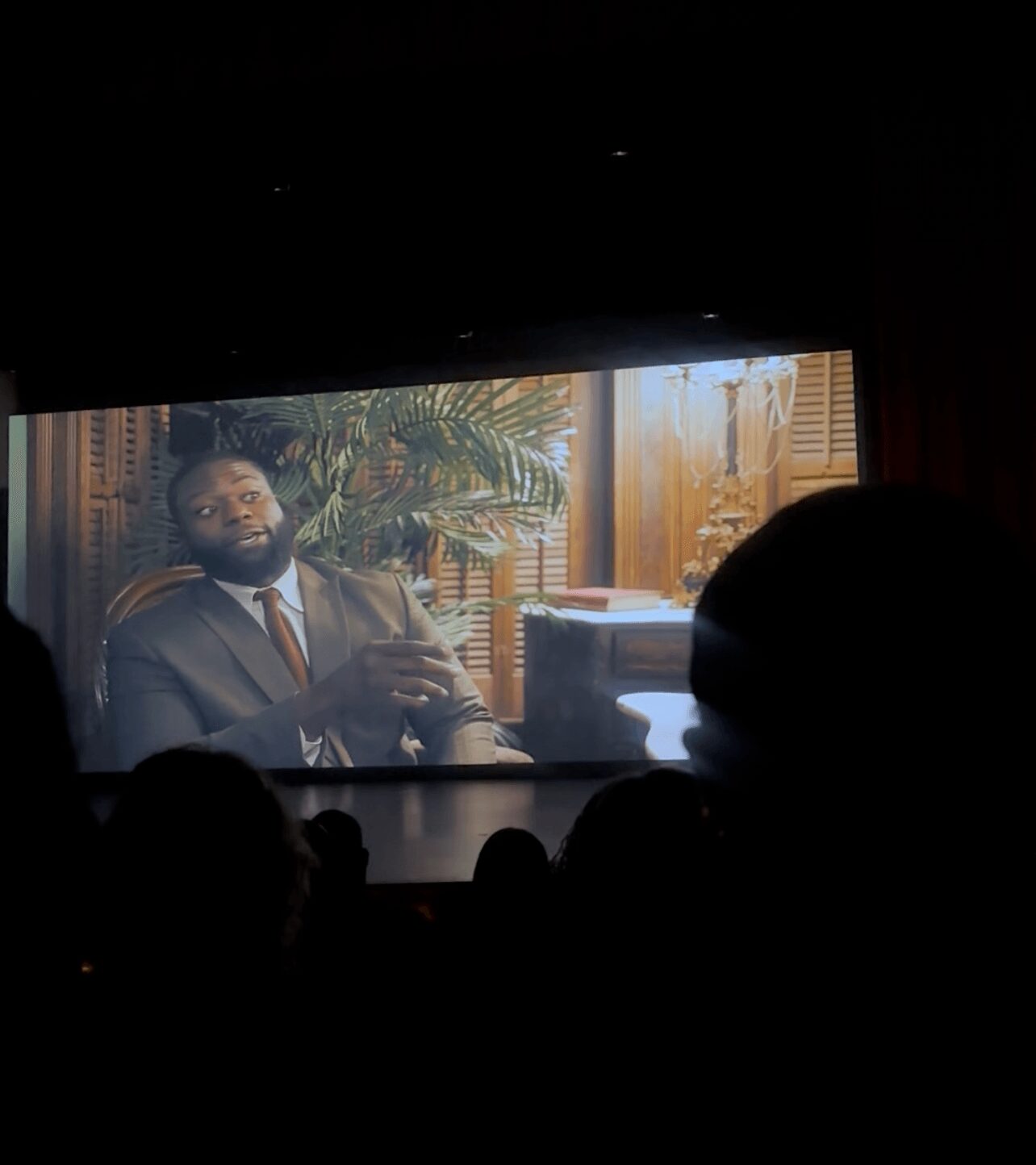 Person in a suit sitting in a room with plants and window blinds, visible on a screen in a dark theater.