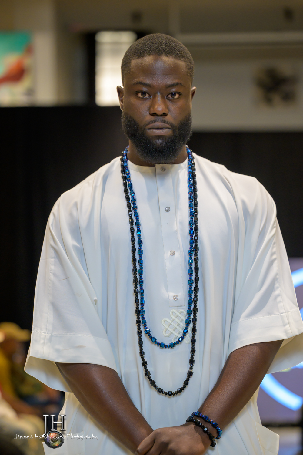 Man with a serious expression wearing white traditional attire and beaded necklaces, standing indoors.