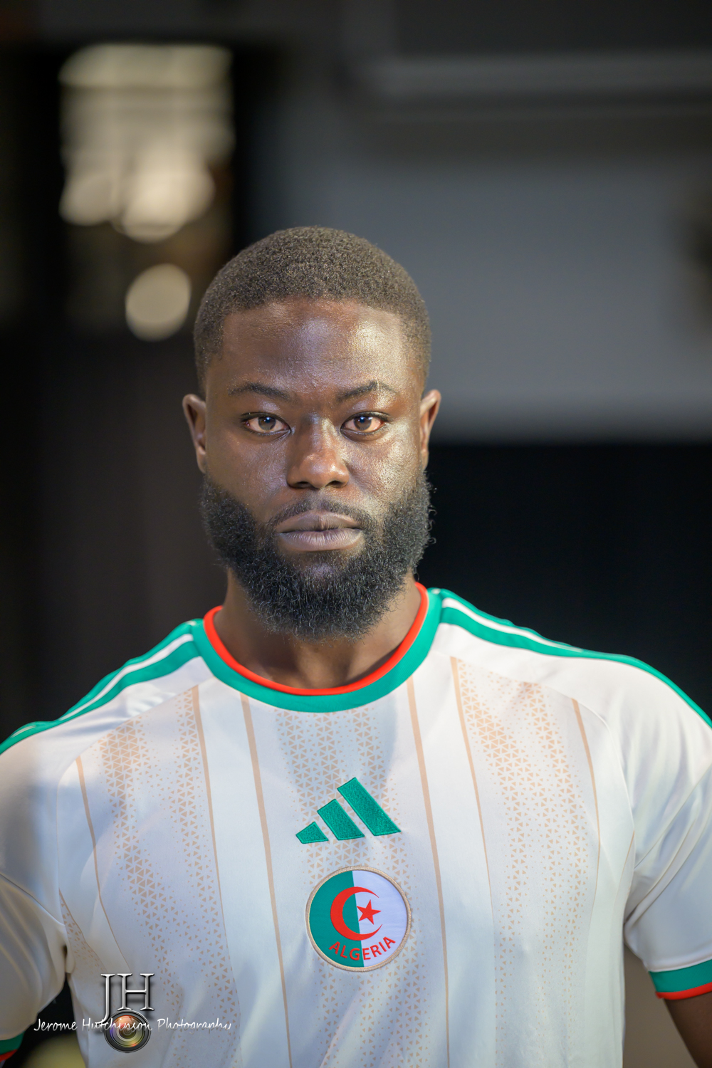 A man with a beard and short hair wearing a white sports jersey with green and red accents, and a logo with a star and crescent moon.