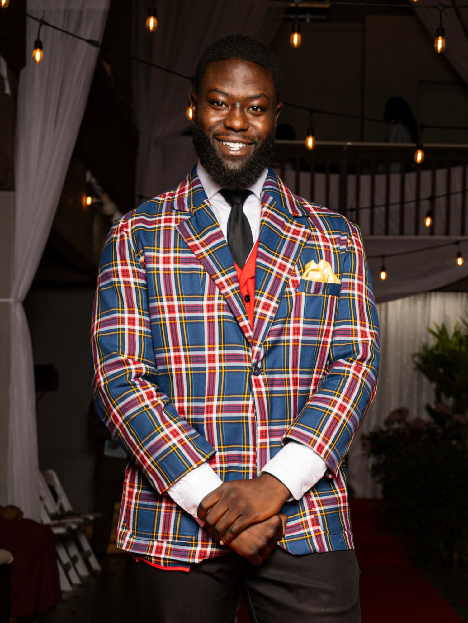 Man in a colorful plaid blazer, white shirt, and tie, smiling indoors with string lights overhead.