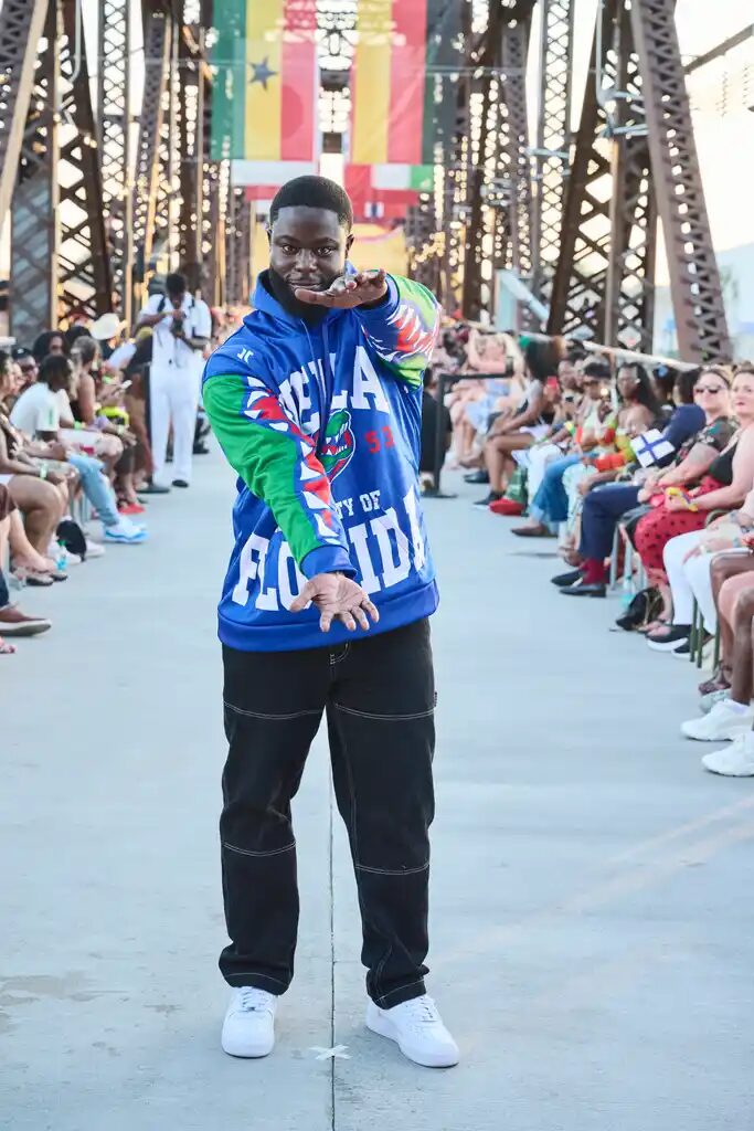 Young person standing on a bridge with colorful flags, surrounded by seated people, smiling and posing with one hand extended.