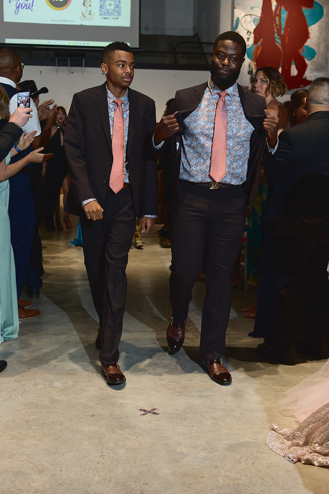 Two men in suits walking and talking at an indoor event, surrounded by other people, some taking photos.