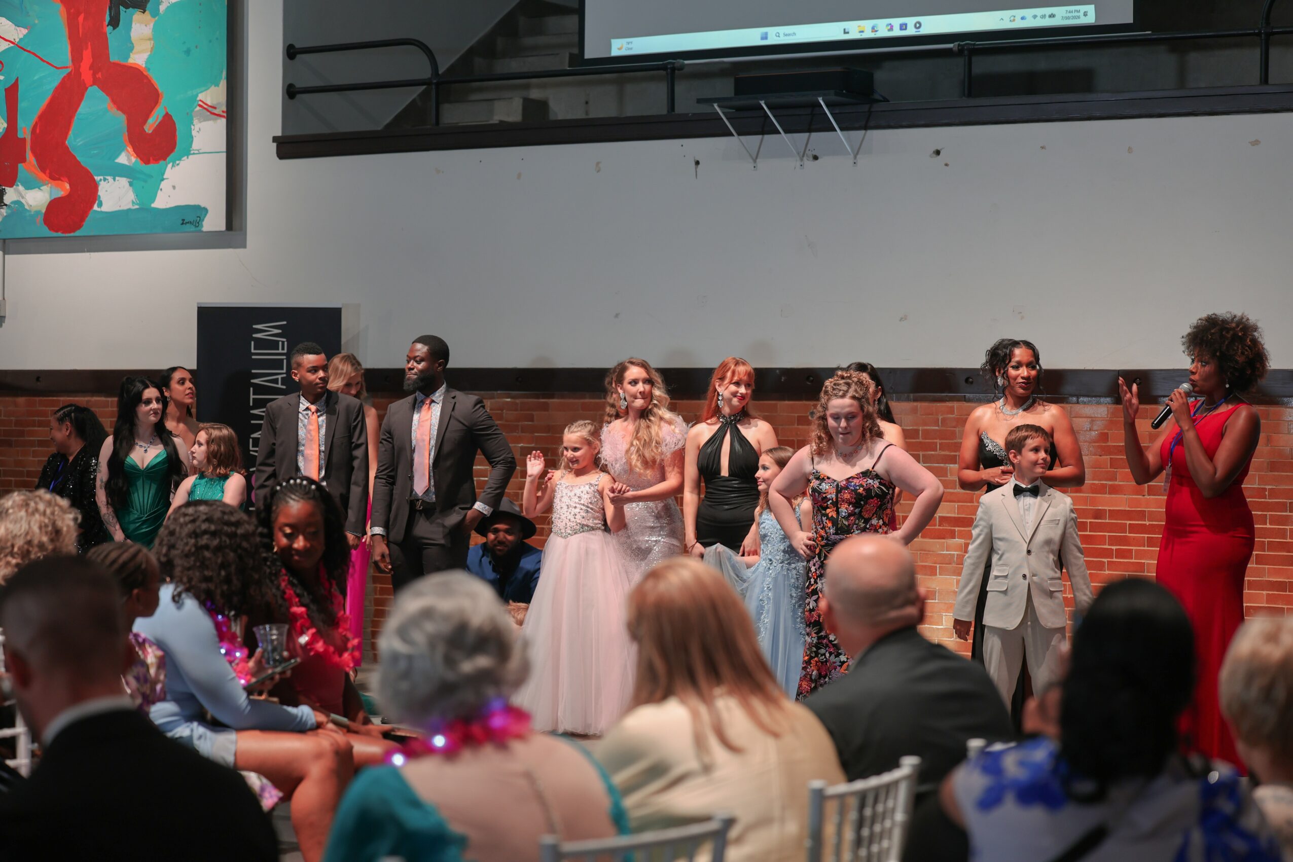 Group of people standing on stage, some in formal attire, with an audience seated in front, in an indoor event space.