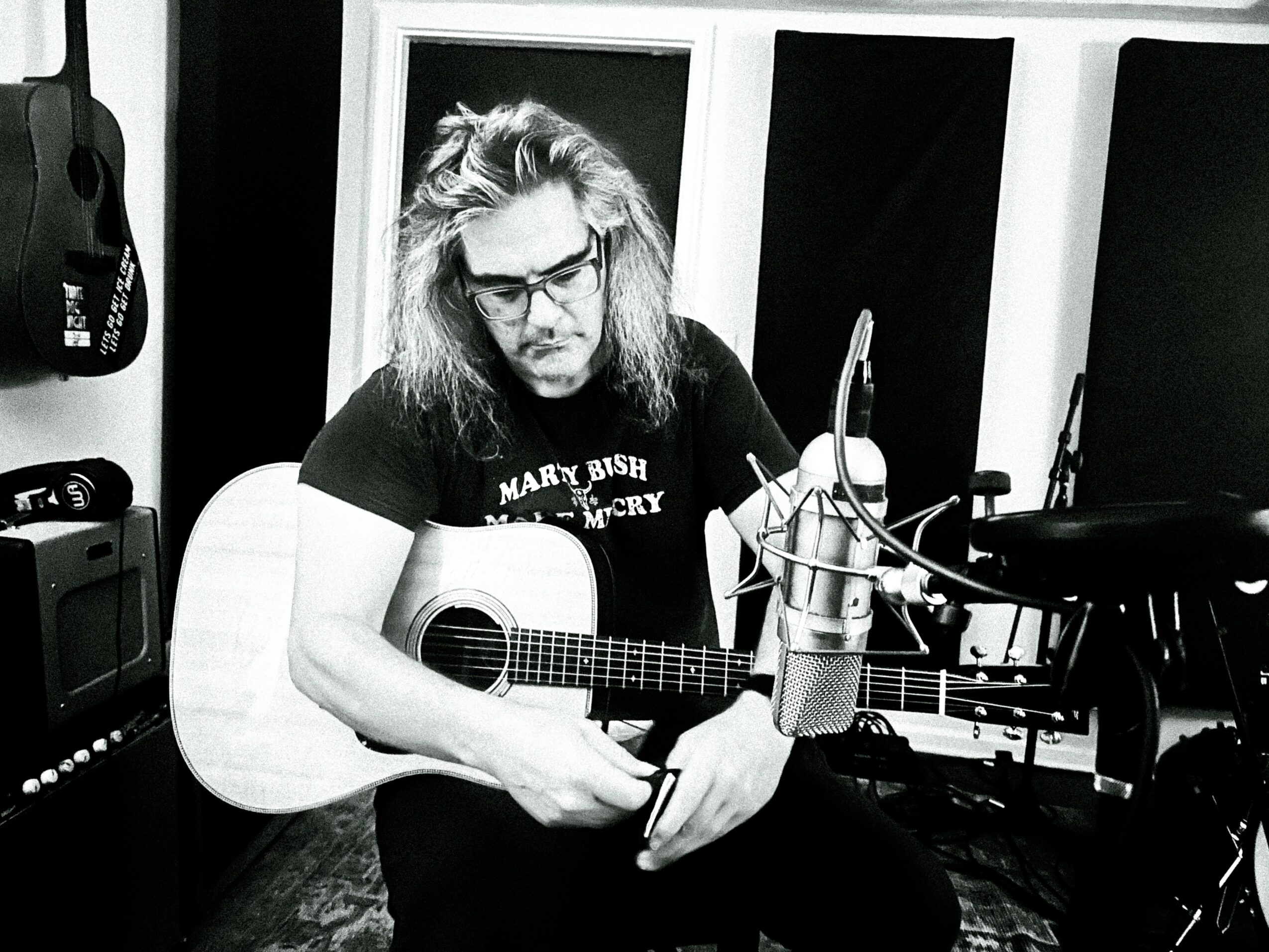 Person with long hair and glasses holding an acoustic guitar in a recording studio, surrounded by equipment.