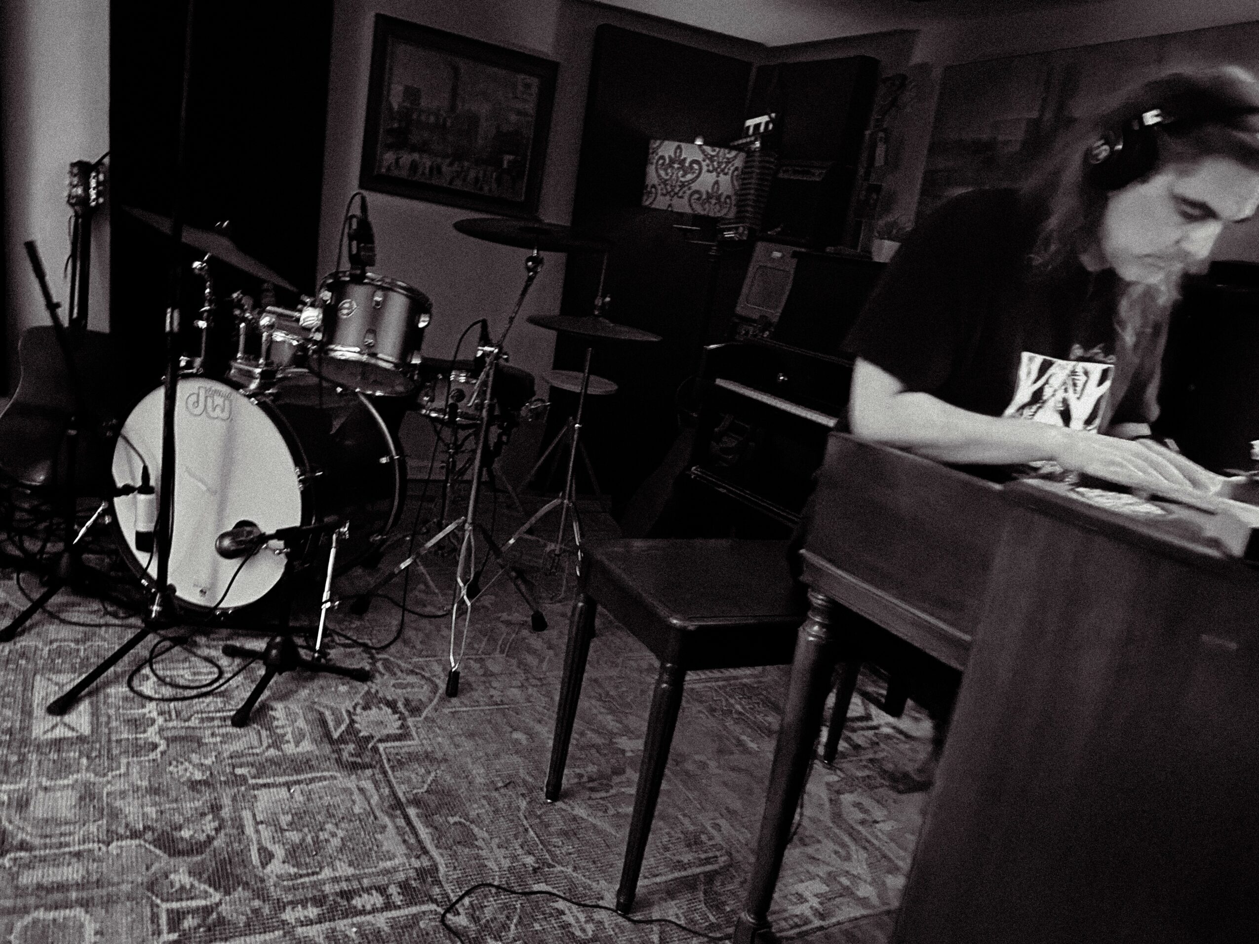 Person playing piano in a room with drums and musical equipment, black and white photo.