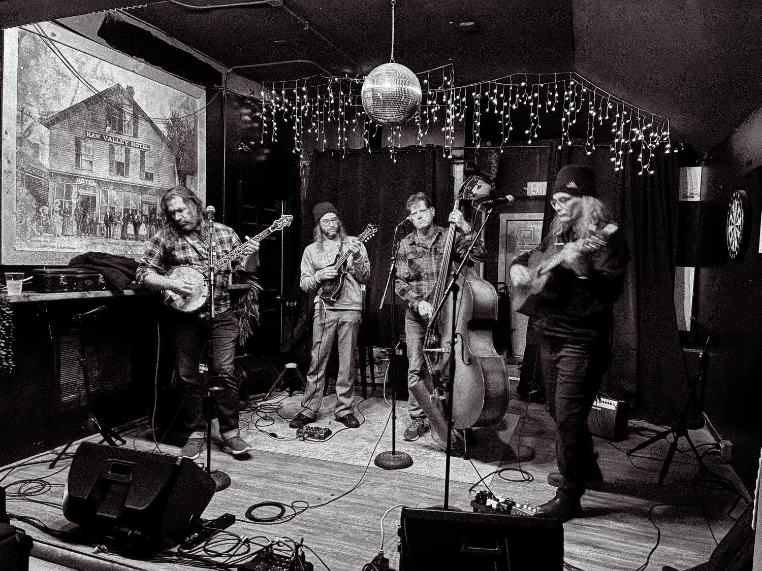 Five musicians perform on stage with instruments, a large painting, and decorative lights in the background.