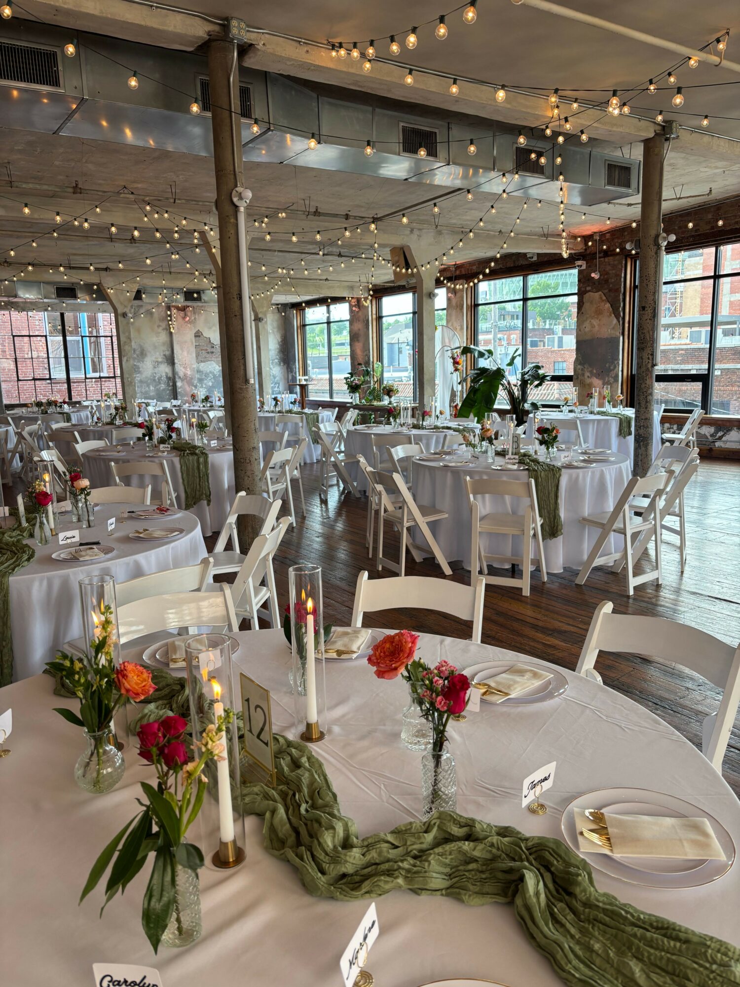 Round table decorated with flowers, candles, and green fabric, set for a celebration in a bright, decorated venue.