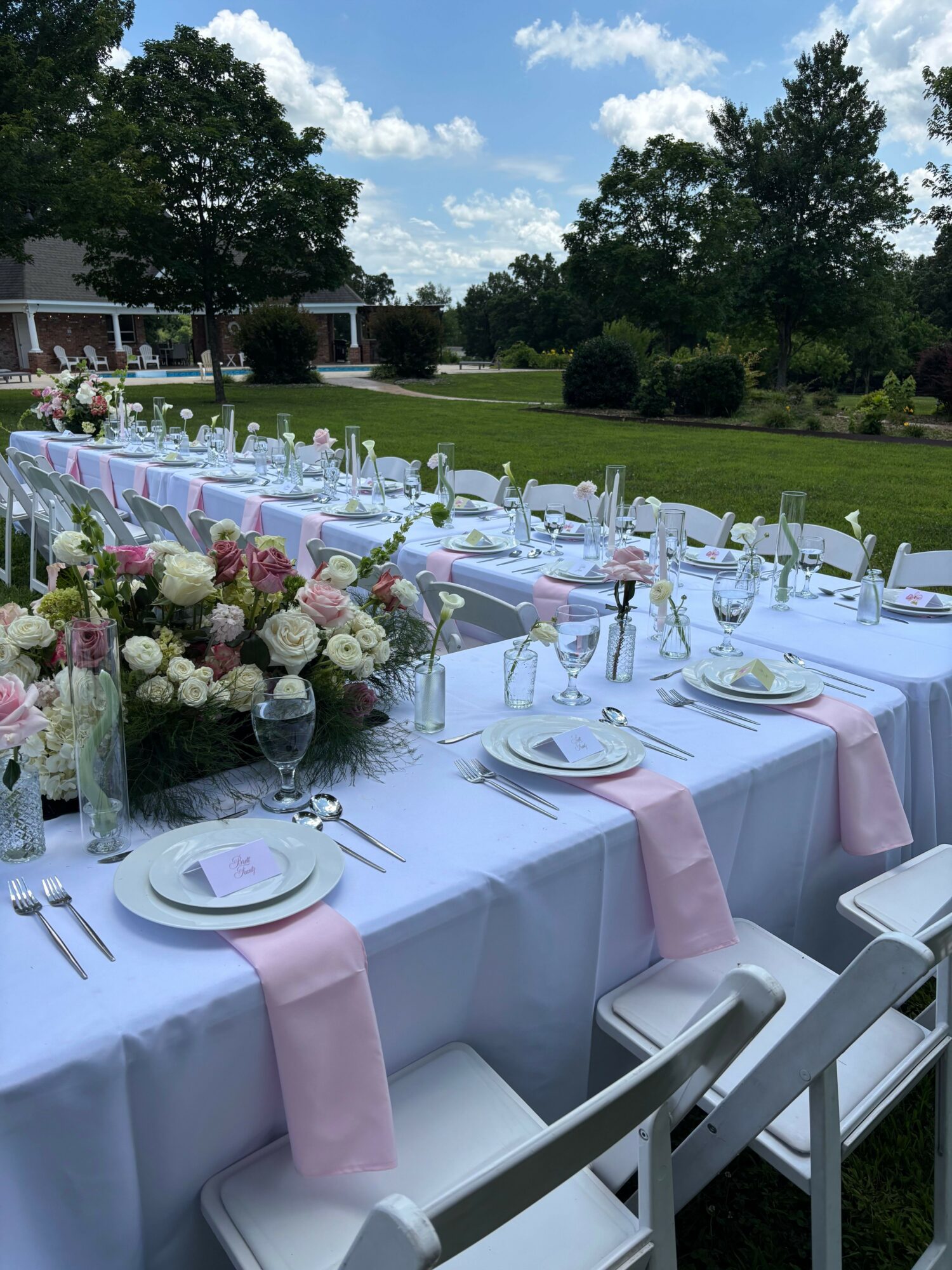 Long outdoor table set with white tablecloth, pink napkins, floral centerpiece, plates, glasses, and cutlery, under trees on a sunny day.