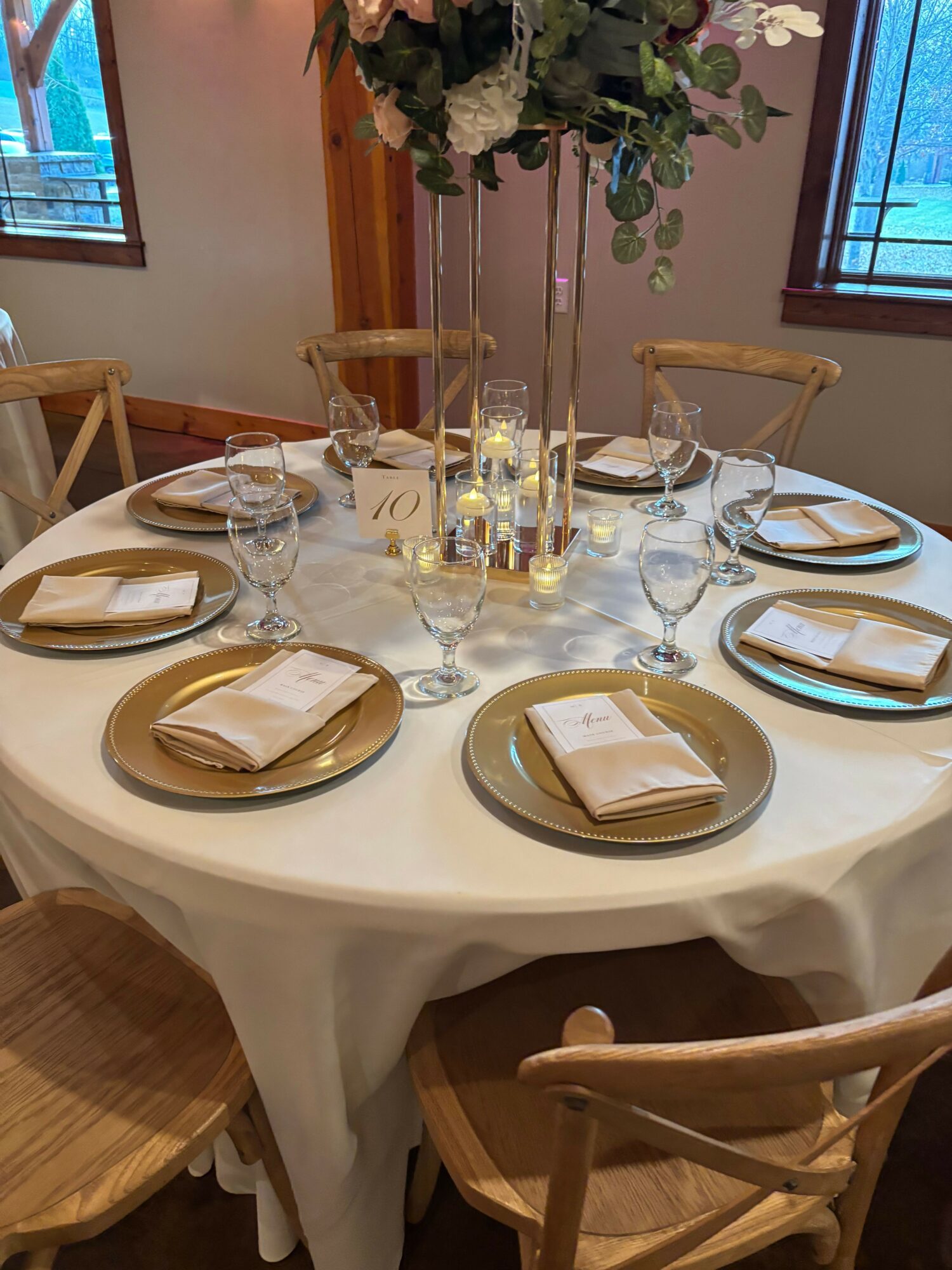Round dining table set with four plates, glasses, and napkins, with chairs around it, in a room with windows and flowers.