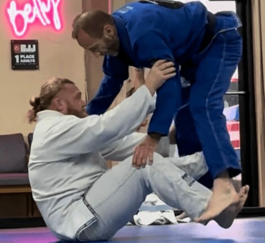 Two women practicing Brazilian Jiu-Jitsu on mats, one in a white gi and the other in a blue gi, engaging in a grappling move.