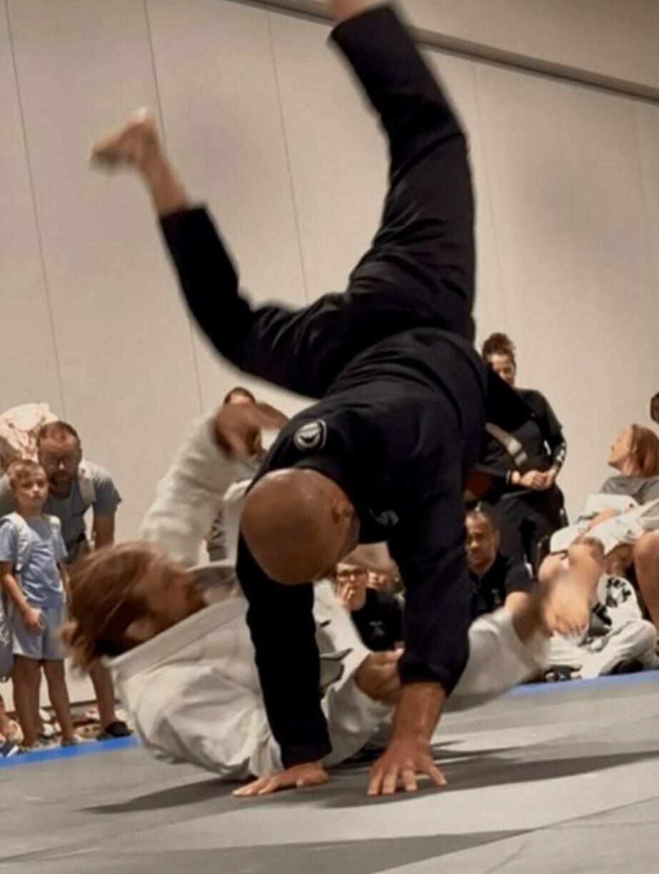 Two people practicing martial arts on mats, one person balancing on hands and feet, others watching in the background.