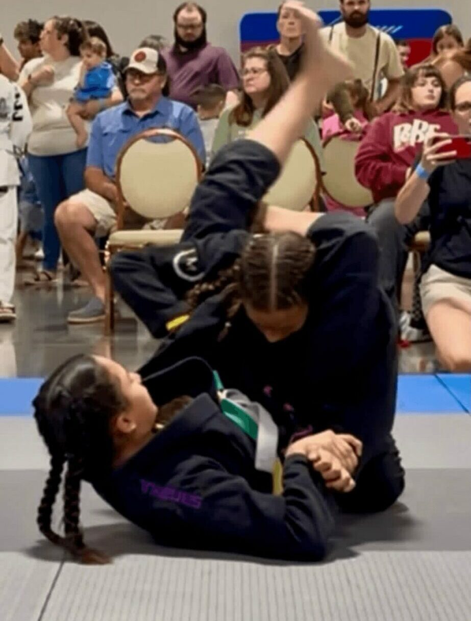 Two women grappling on the floor at a competition, with spectators watching in the background.
