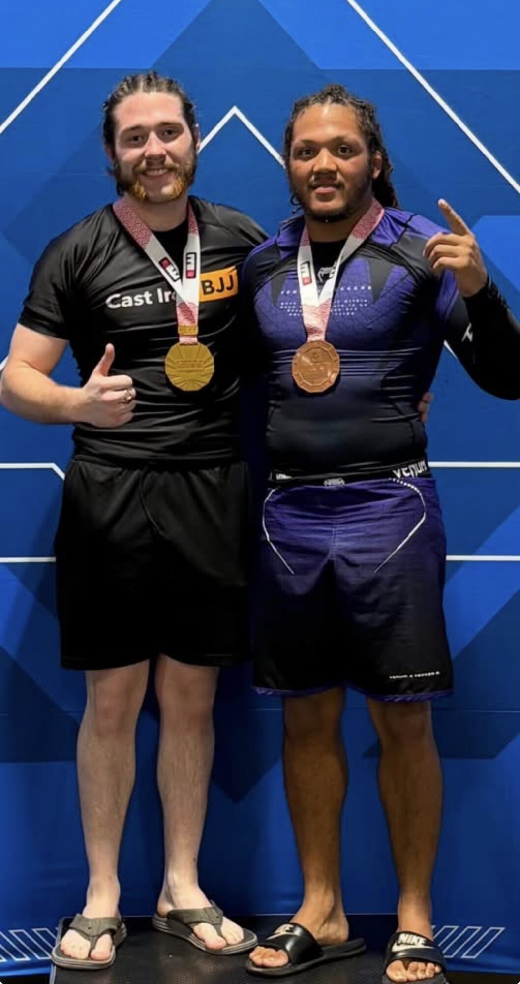 Two male athletes with medals, standing side by side, smiling, in front of a blue geometric background.