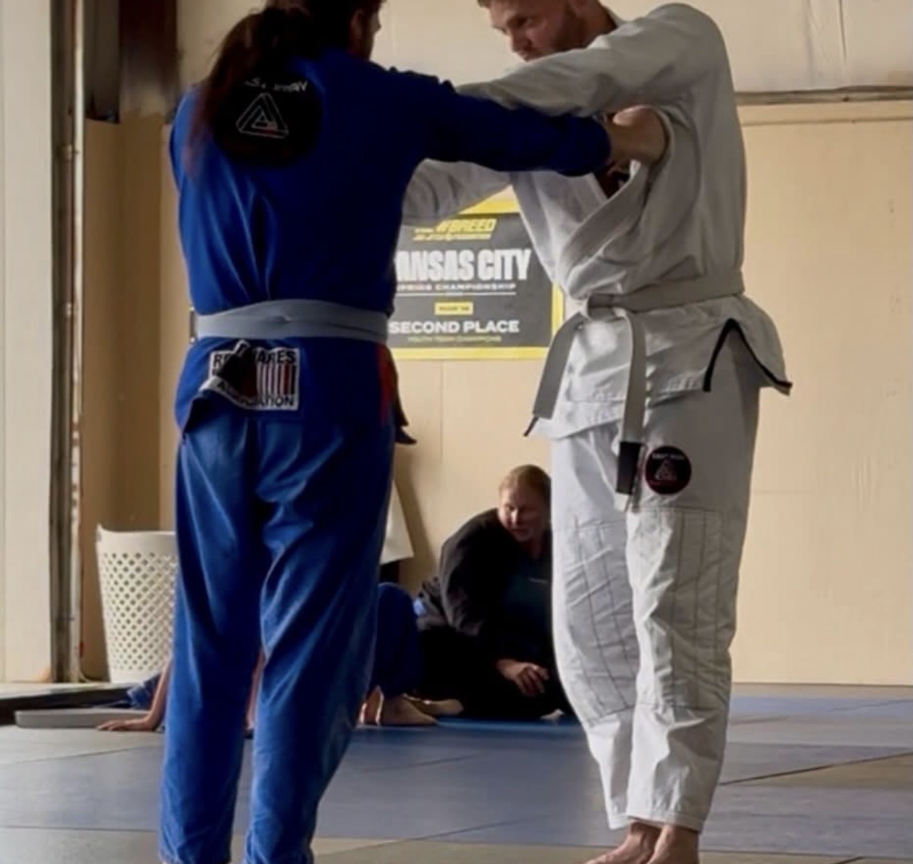 Two people in martial arts uniforms practicing techniques in a dojo, with a person sitting on the floor in the background.