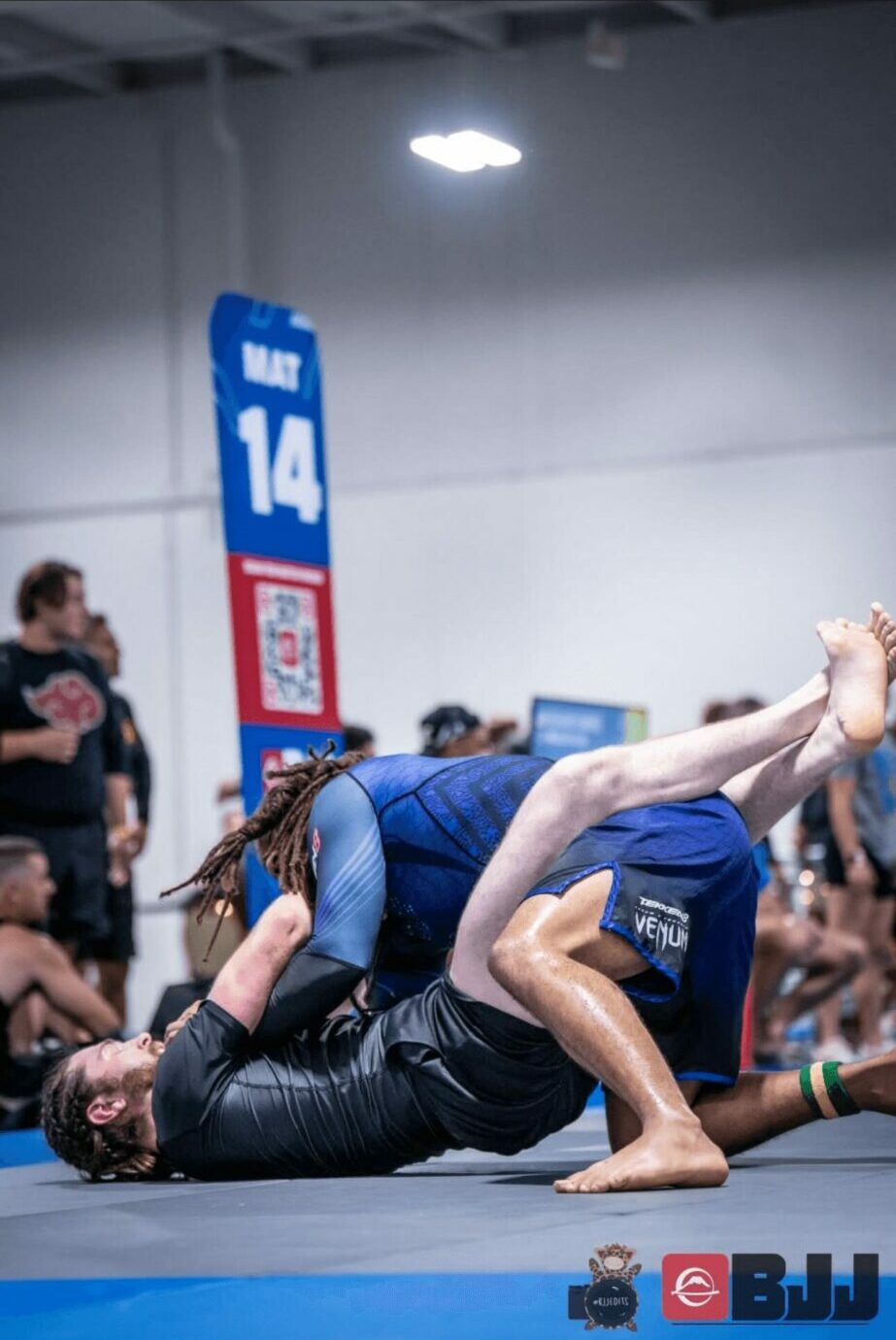 Two women compete in a Brazilian Jiu-Jitsu match on blue mats, with spectators watching in the background.