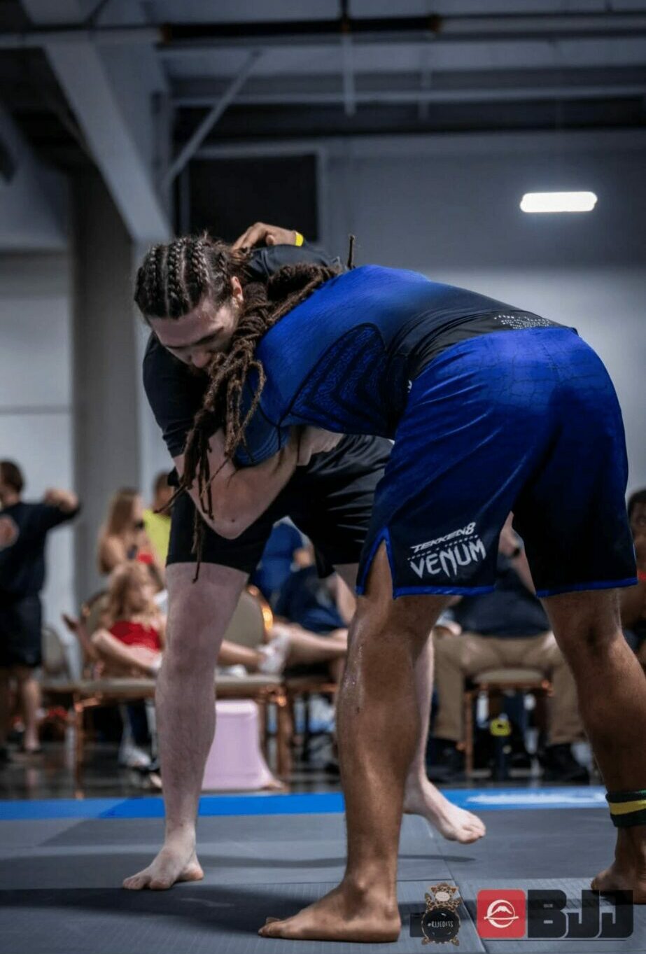 Two people practicing Brazilian Jiu-Jitsu on mats in a gym, with others in the background. One person is in a dominant position.