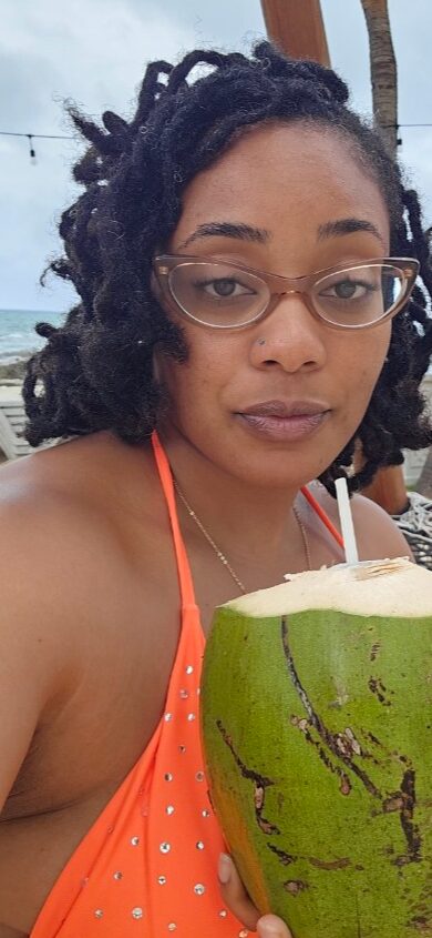 Woman with glasses and dreadlocks holding a green coconut with a straw, outdoors, wearing an orange top.