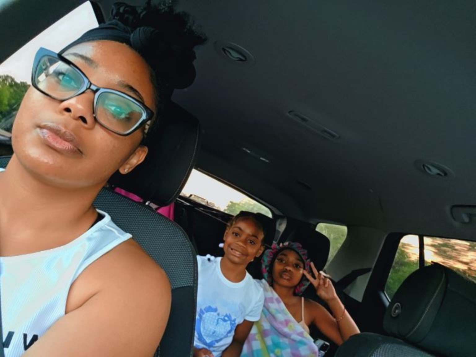 Three women sitting in a car, two making peace signs, one wearing glasses, all smiling.