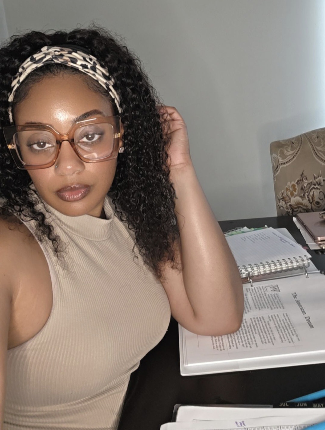 Woman with curly hair, glasses, and a headband, sitting at a desk with open books and papers.
