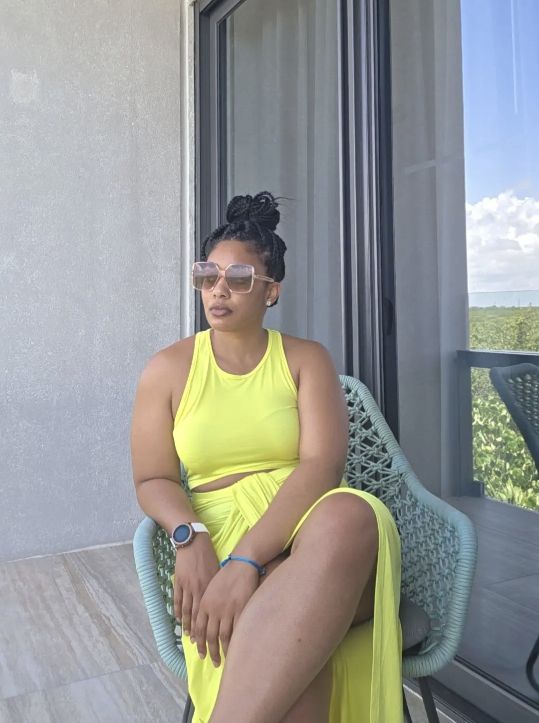 Woman in a yellow dress sitting on a chair near a large window with a view of blue sky and greenery.