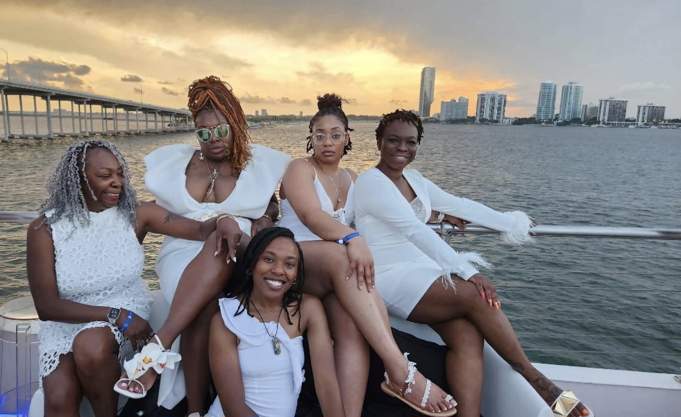Five women sitting on a boat with a city skyline and water in the background, smiling and enjoying the view.