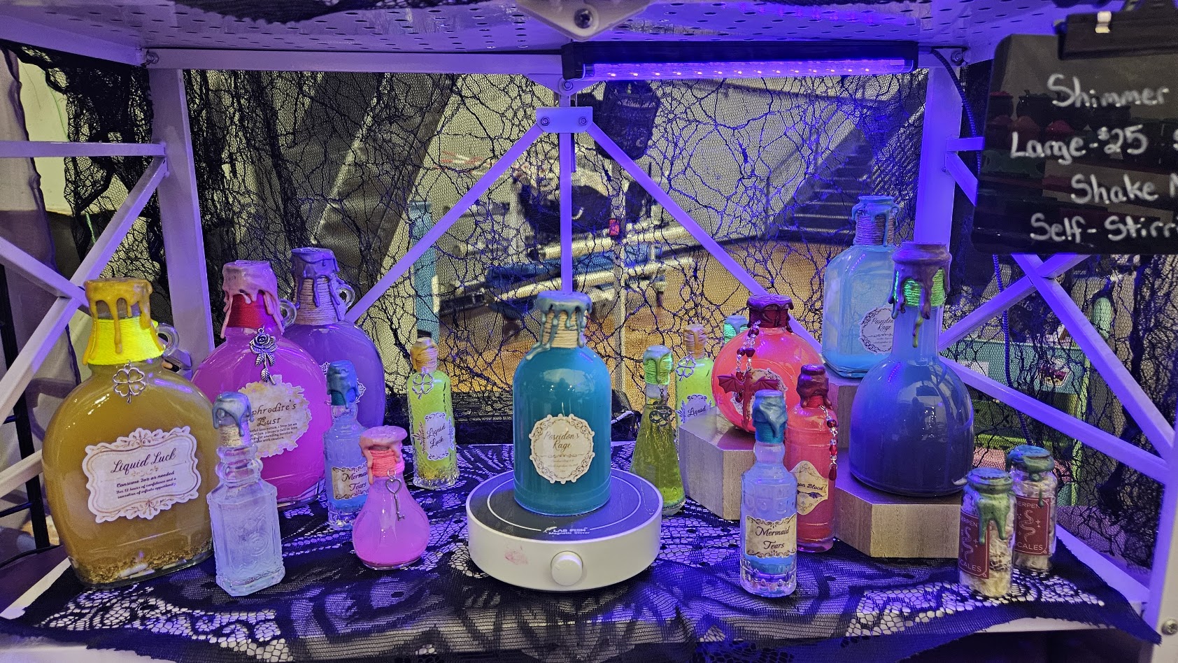 Display of colorful bottles and containers on a table with a black cloth, behind a wire fence, with a chalkboard sign.