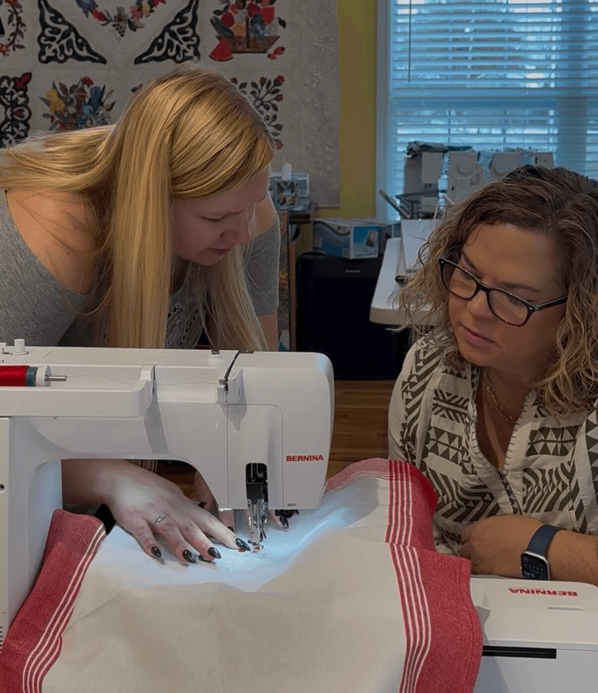 Two women work together on a sewing machine, one guiding fabric and the other observing closely in a room with window blinds.
