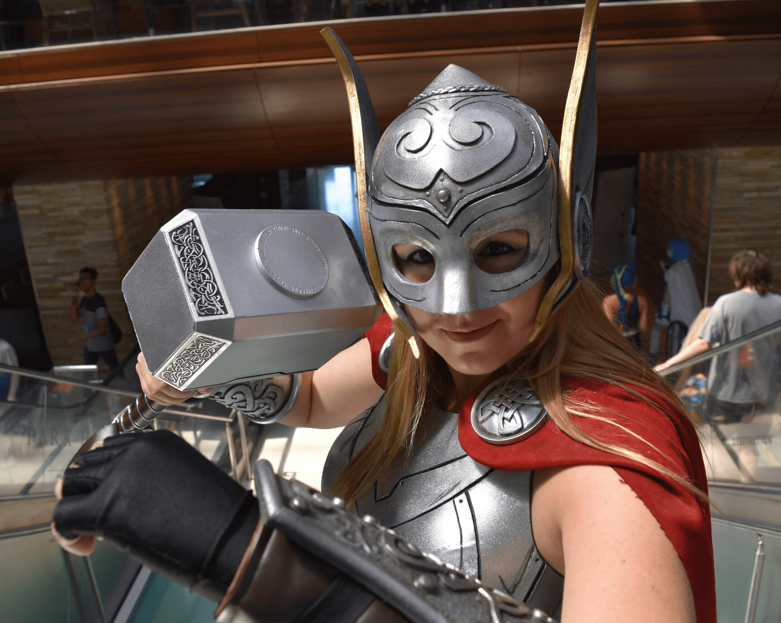 Person in Thor costume with helmet, red cape, and hammer, posing at an indoor event, smiling at the camera.