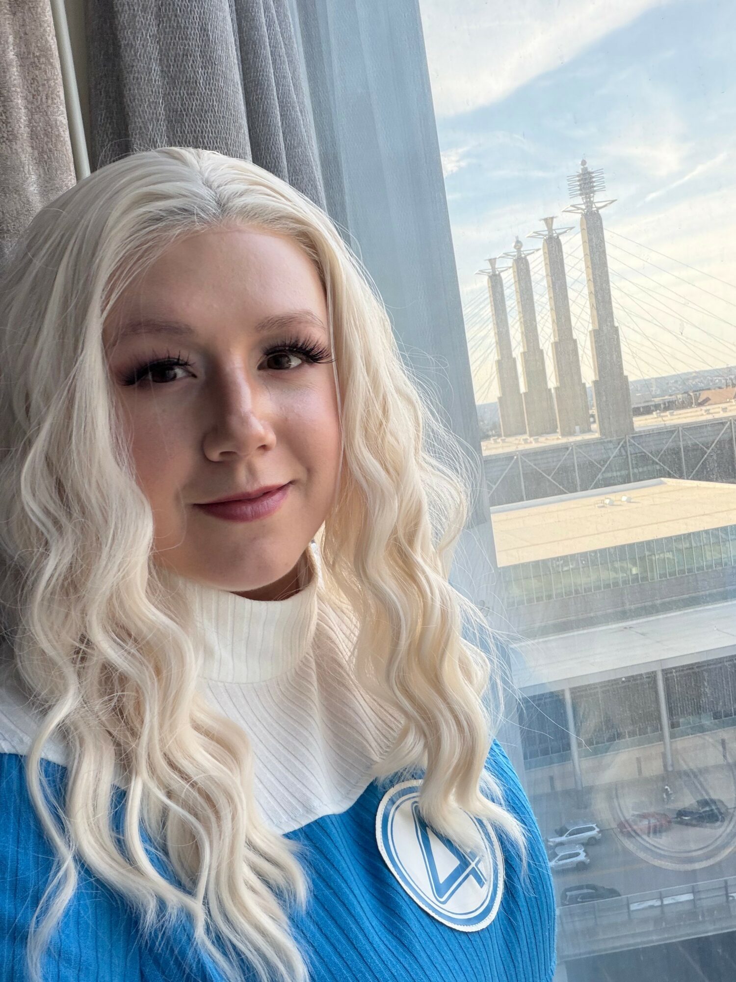 Young woman with long, wavy blonde hair smiling near window with cityscape and bridge in background.