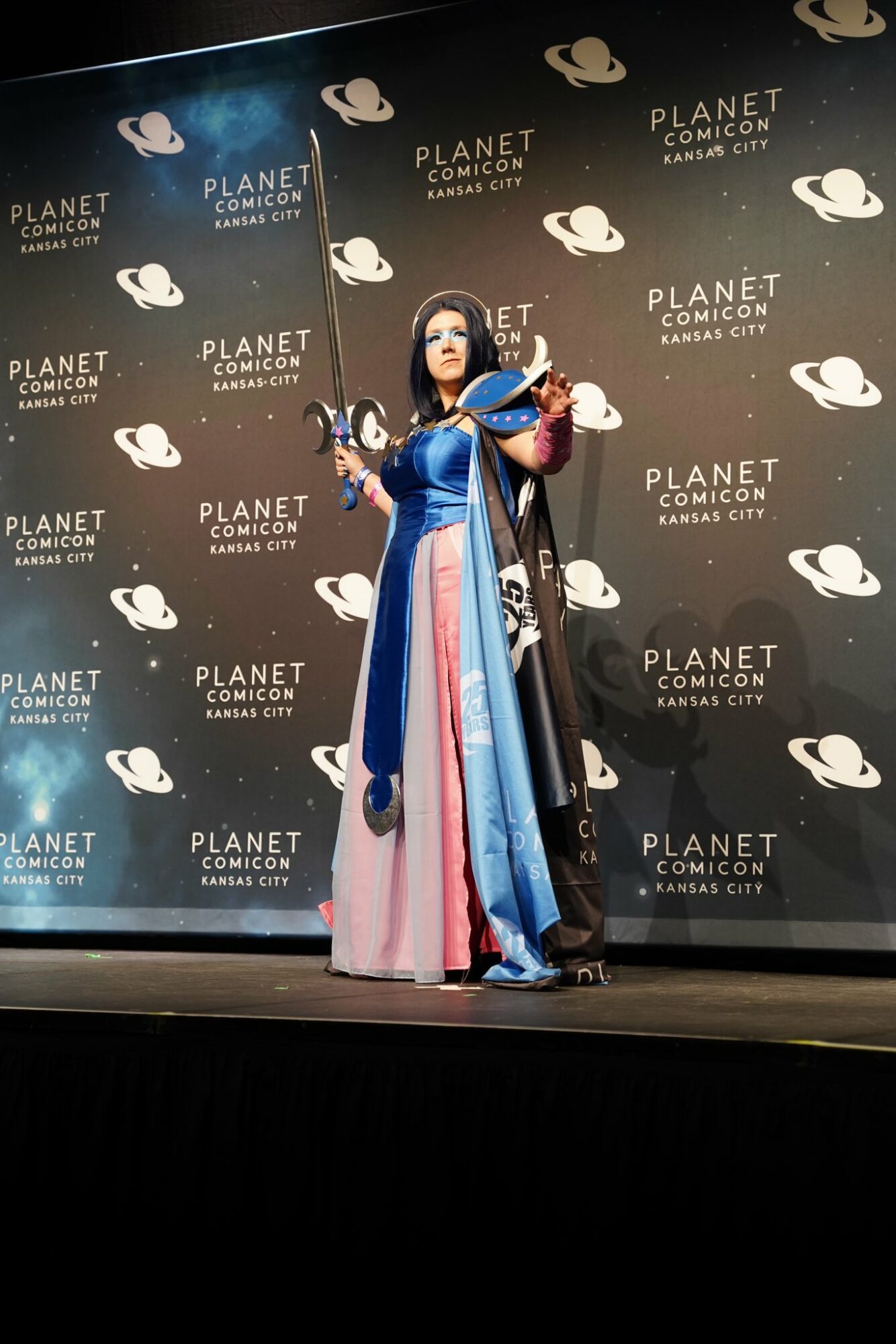Person in colorful costume on stage with a black backdrop featuring white logos and text, holding a microphone.