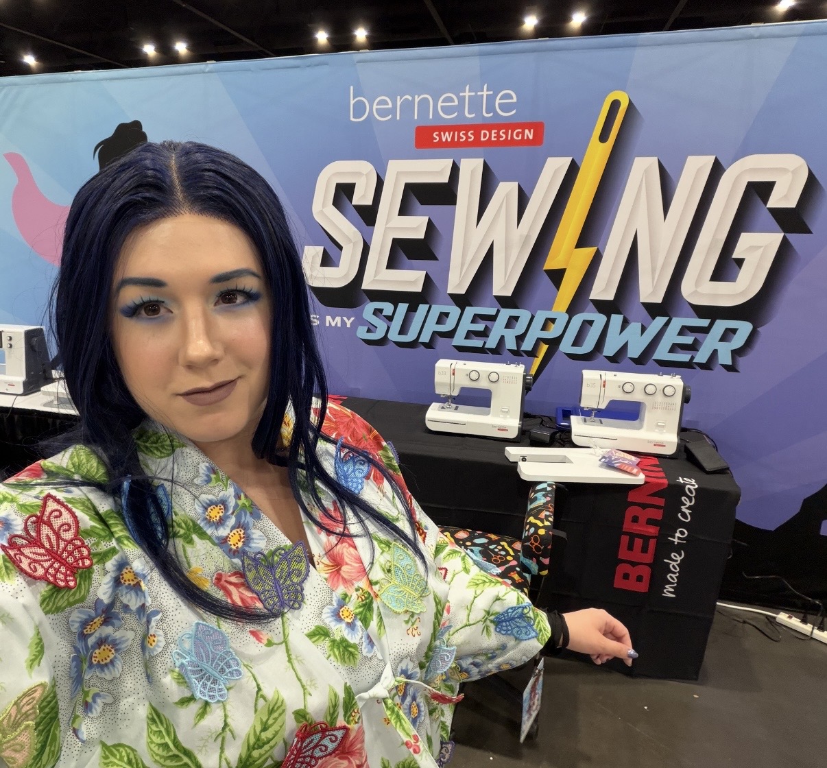 Woman with dark hair in colorful floral top standing in front of sewing machine display at event.