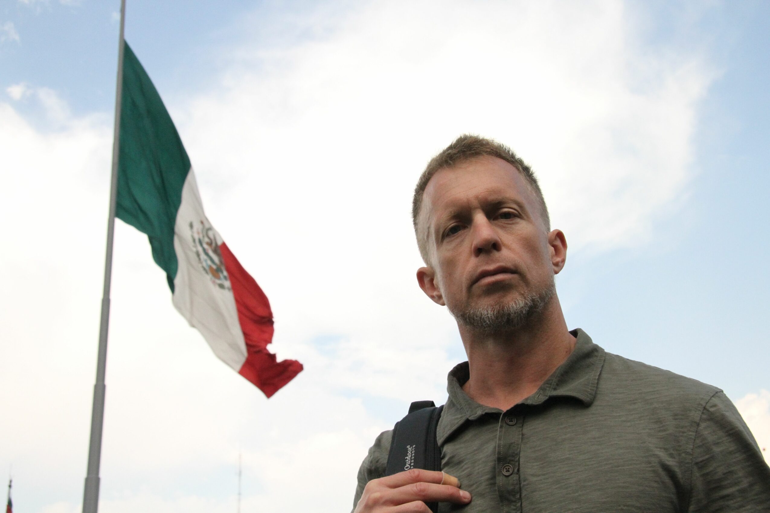 Man with short hair and beard standing outdoors with a flag in the background, sky with clouds, wearing a dark shirt and backpack.