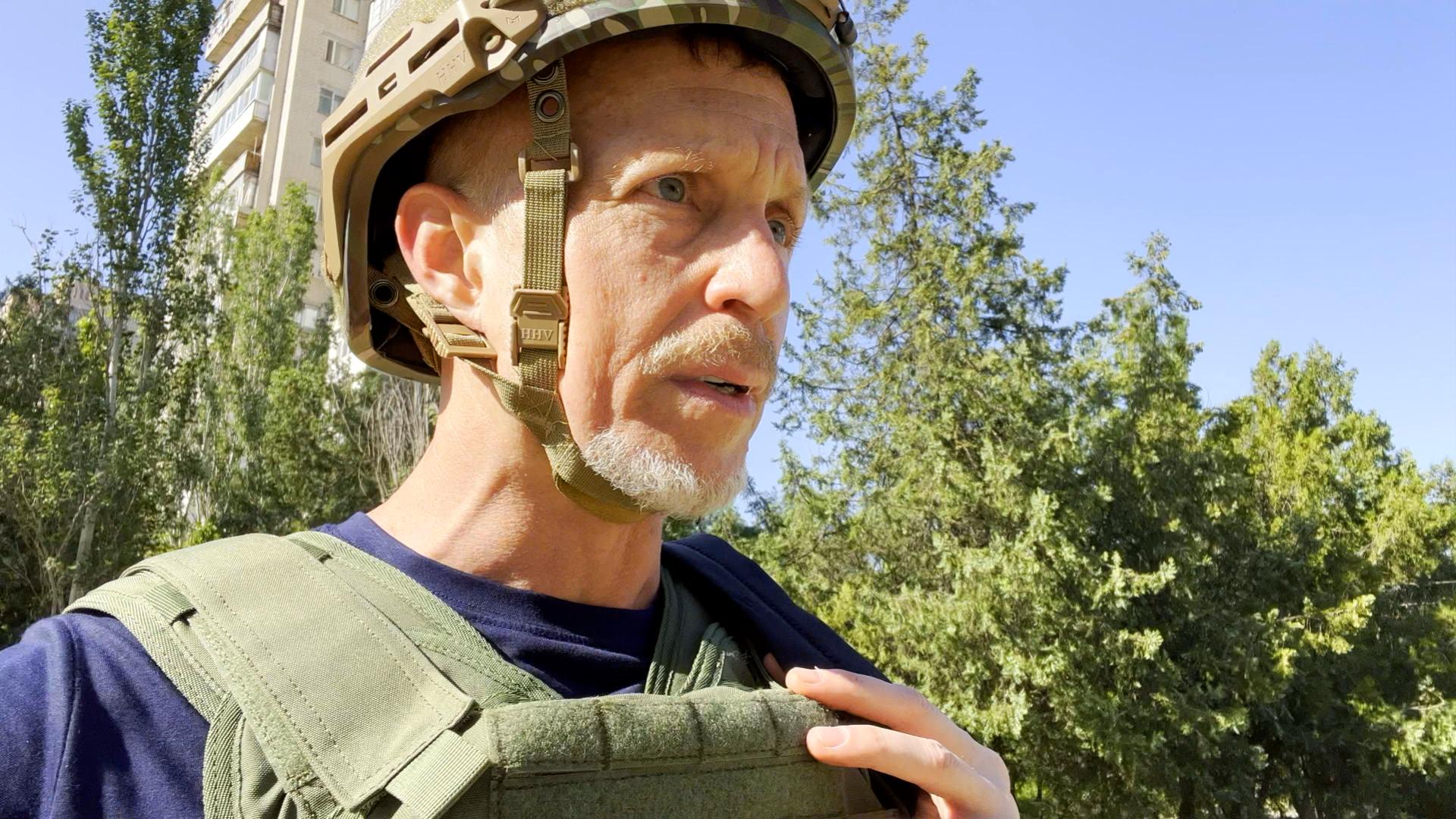 Man wearing military helmet and vest outdoors with trees and blue sky in background.