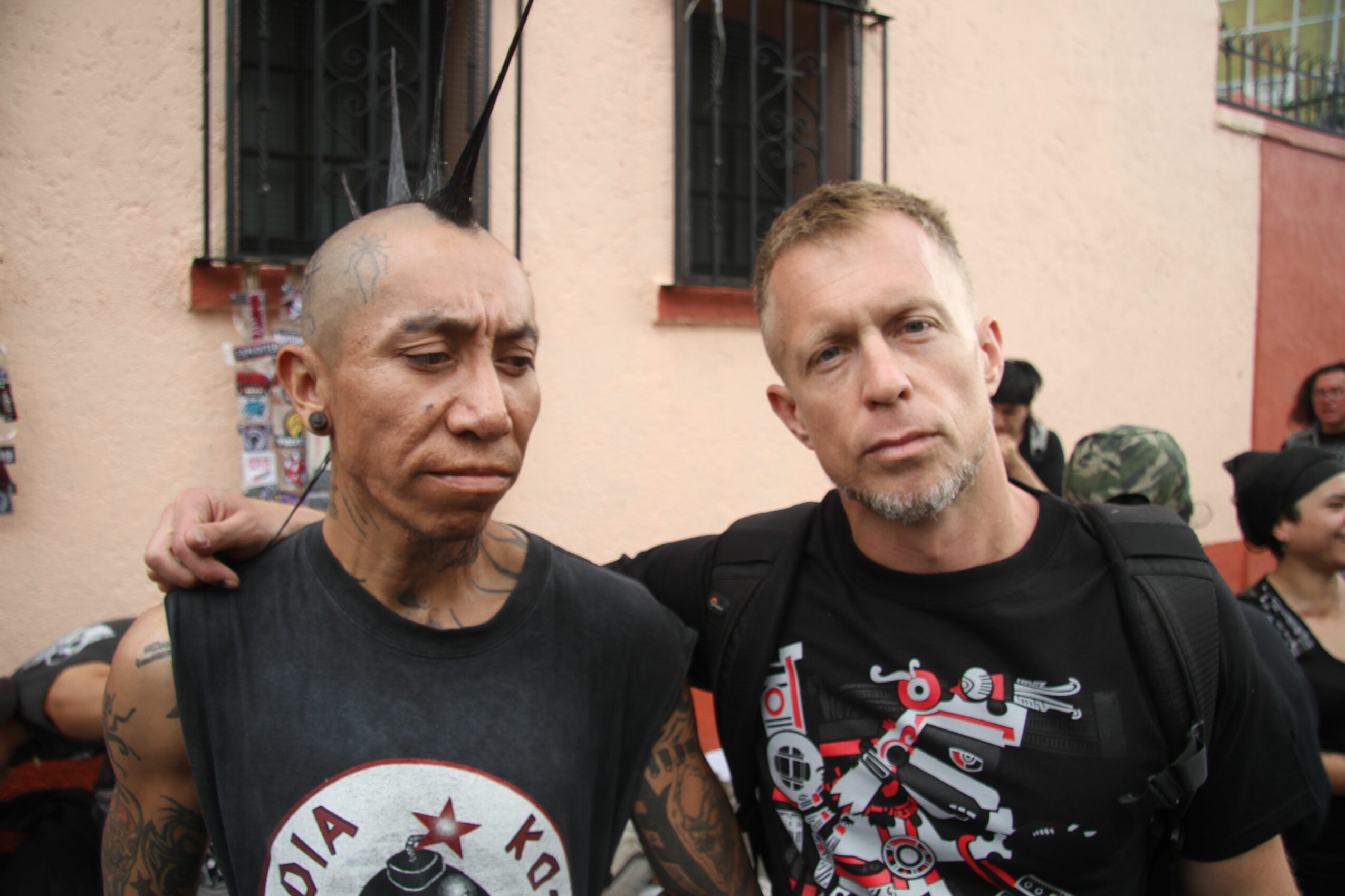 Two men standing close together outdoors, one with a shaved head and the other with short hair, both wearing black shirts.