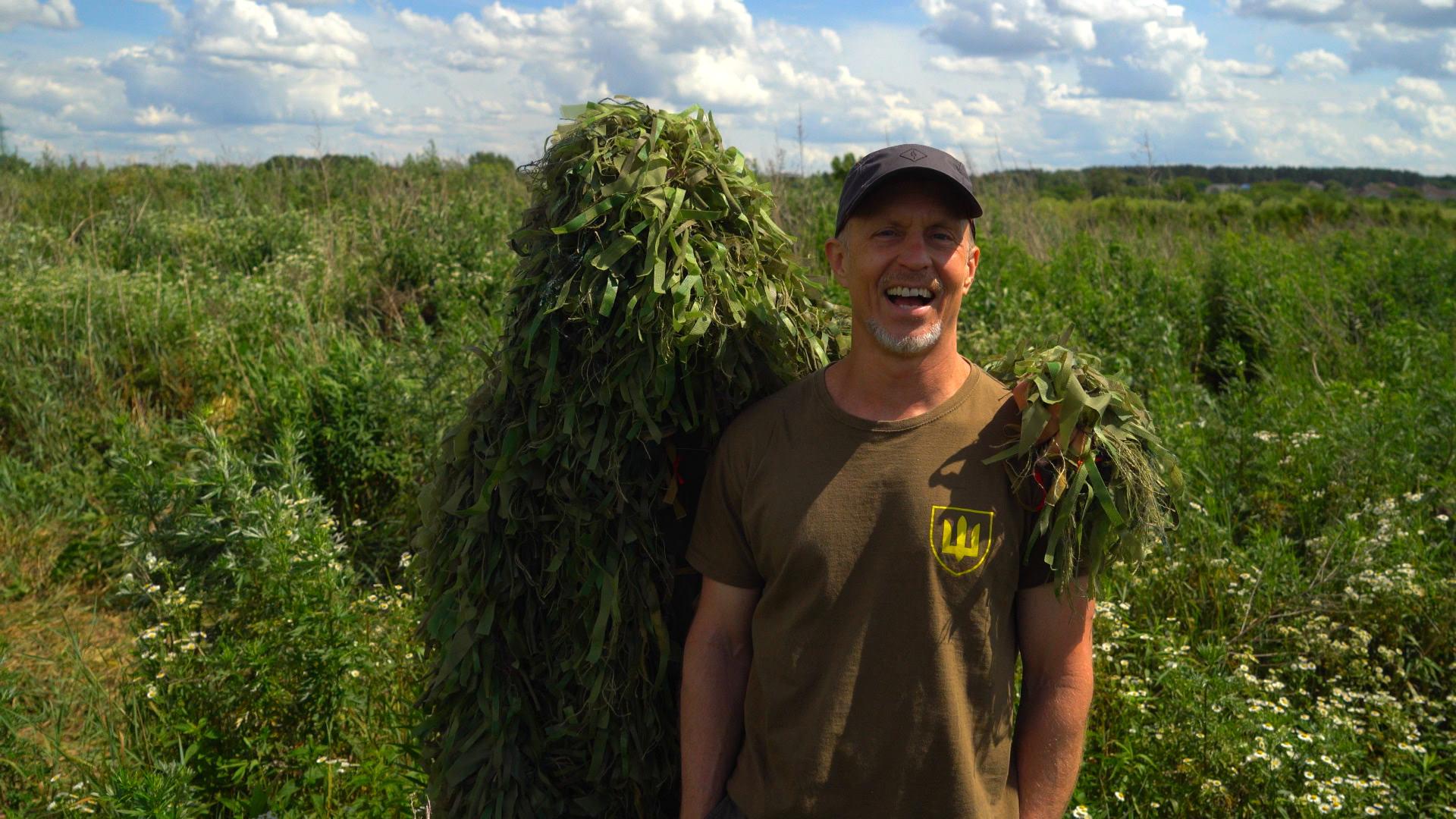 Man smiling outdoors with a large plant on his shoulder, surrounded by green fields and a partly cloudy sky.